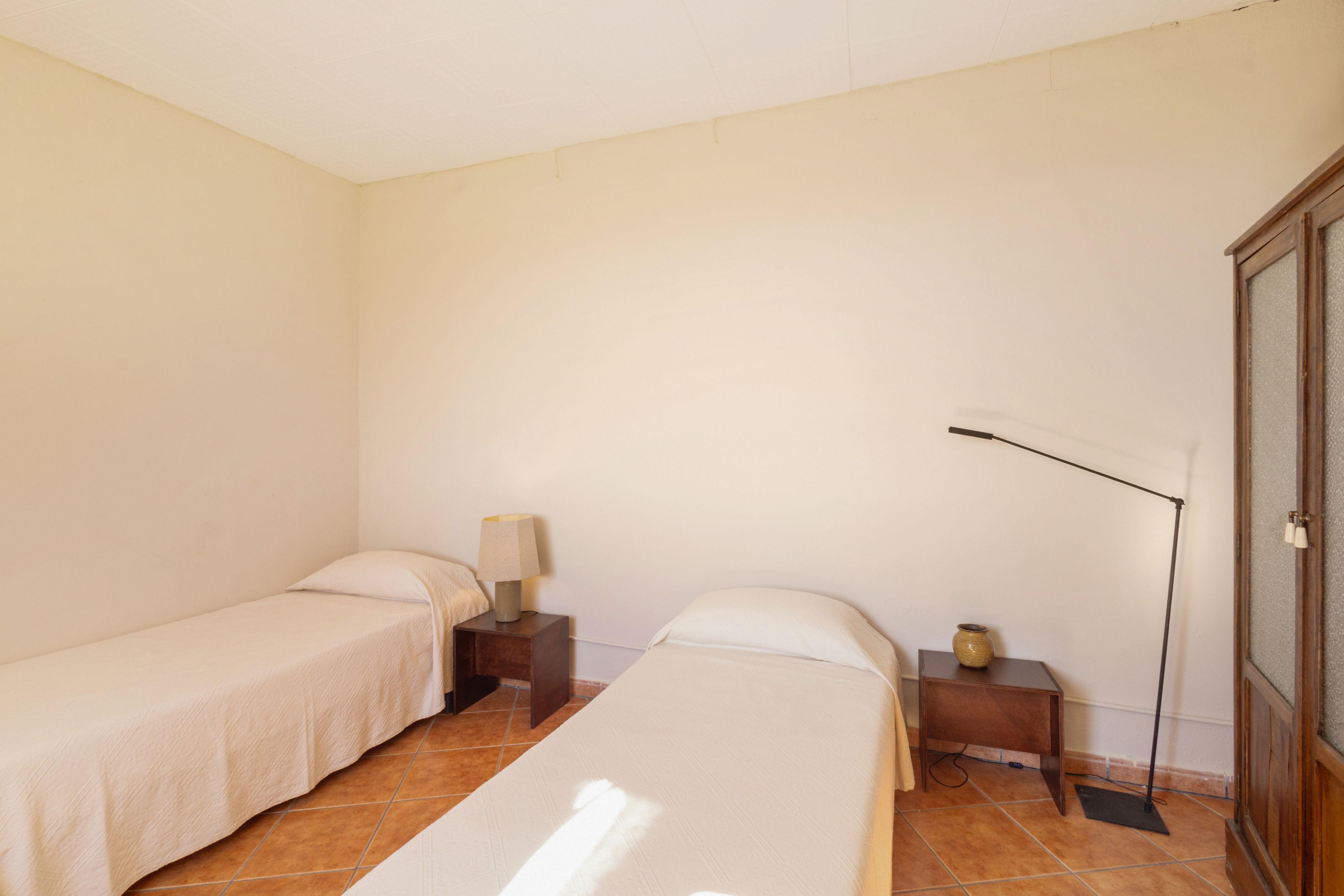 A small, clean room with two single beds, a nightstand with a lamp, and a wardrobe.