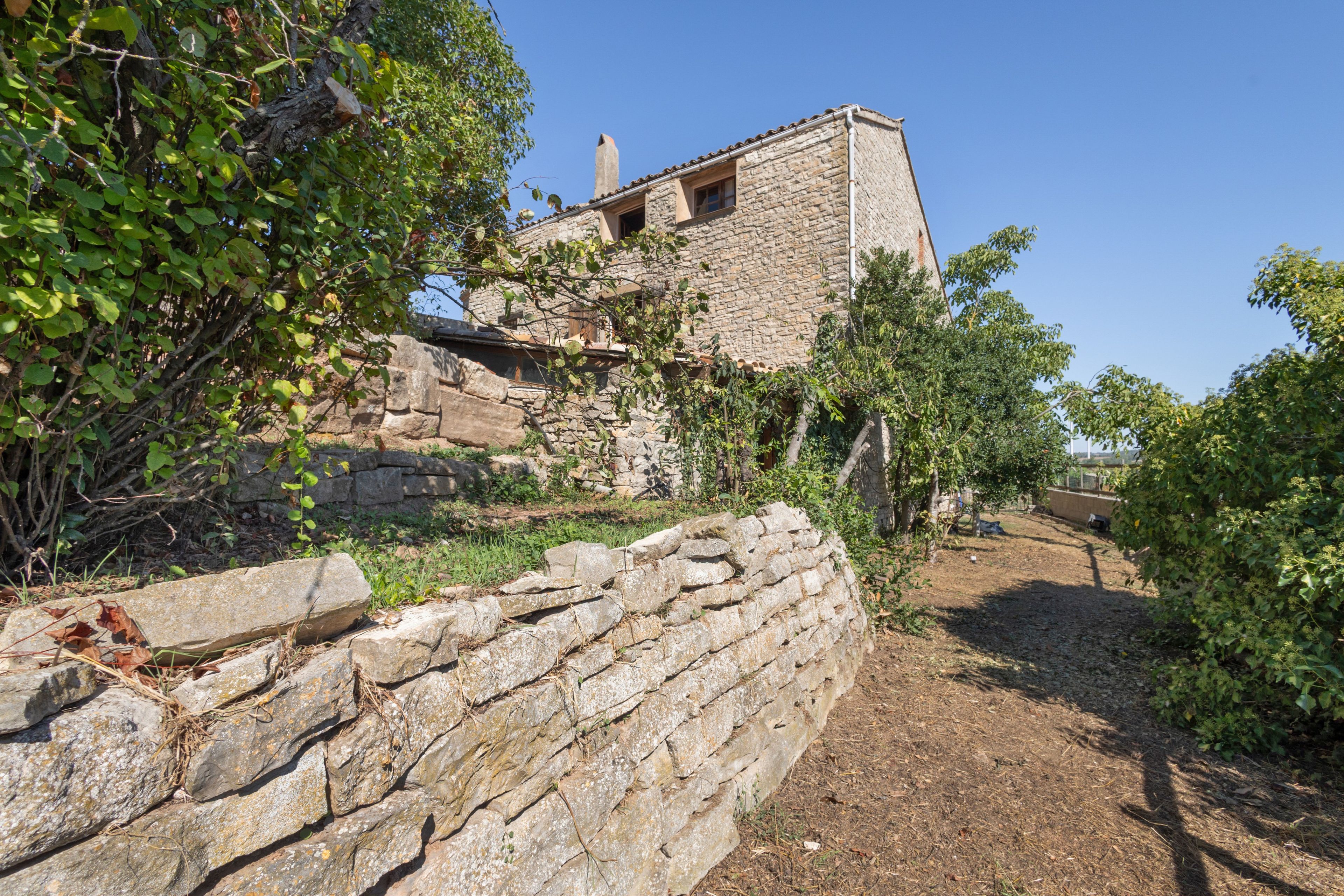 Un edificio de piedra con aspecto rústico, rodeado de vegetación y un muro de piedra.