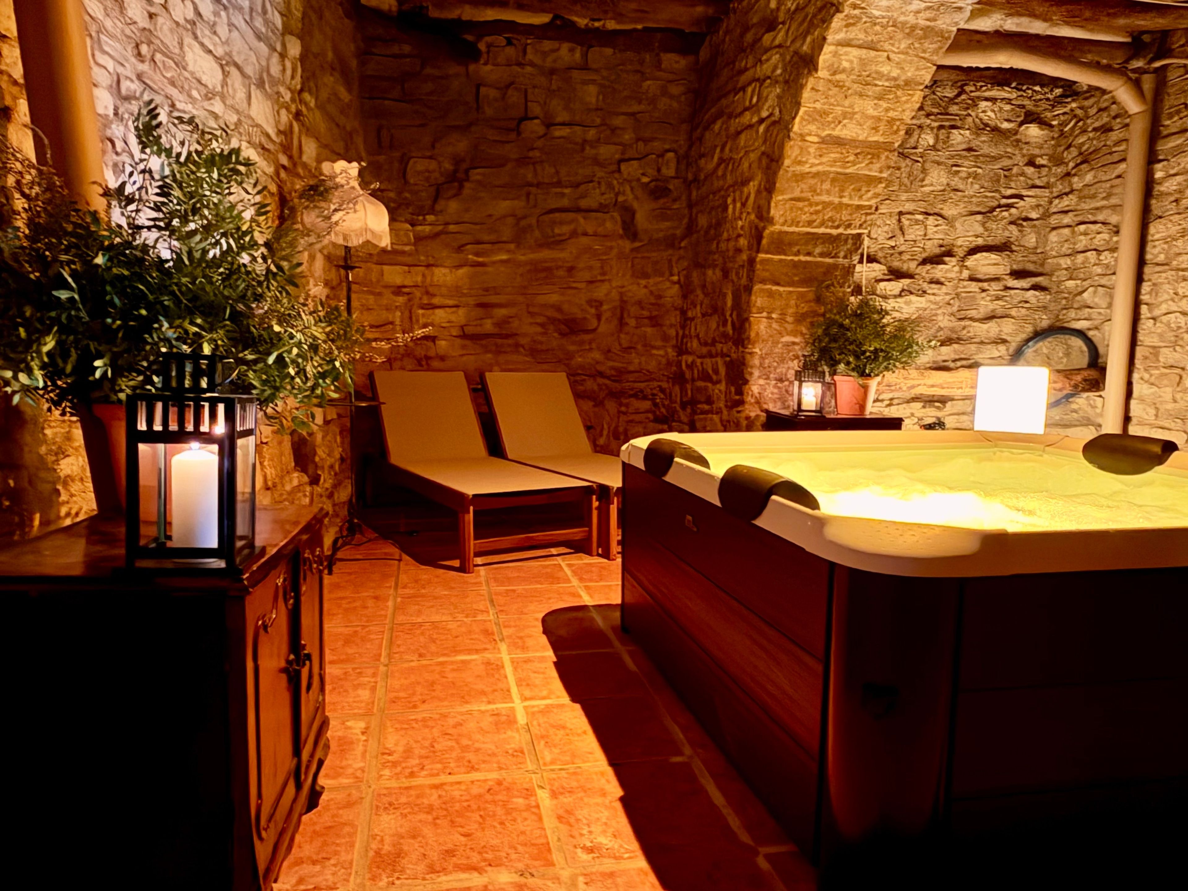 Habitación de paredes de piedra con un jacuzzi, dos sillones de descanso, un farol y plantas en macetas.