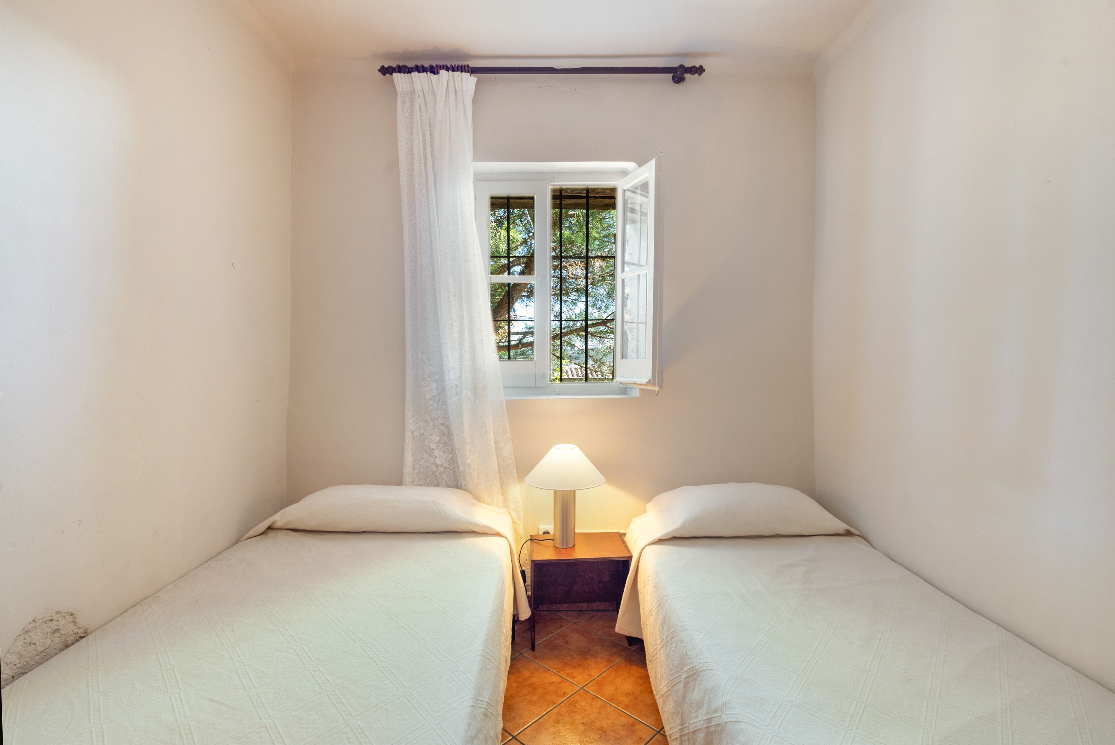 Un alojamiento pequeño, de una sola habitación, con dos camas, un ventanal con vistas a árboles y una lámpara de noche.