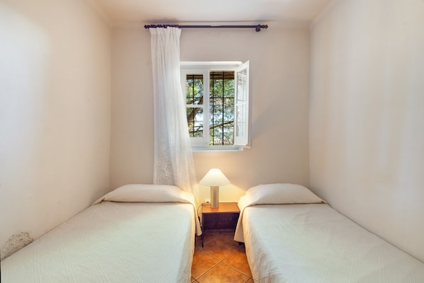 A small, single-room accommodation with two beds, a window with a view of trees, and a bedside lamp.