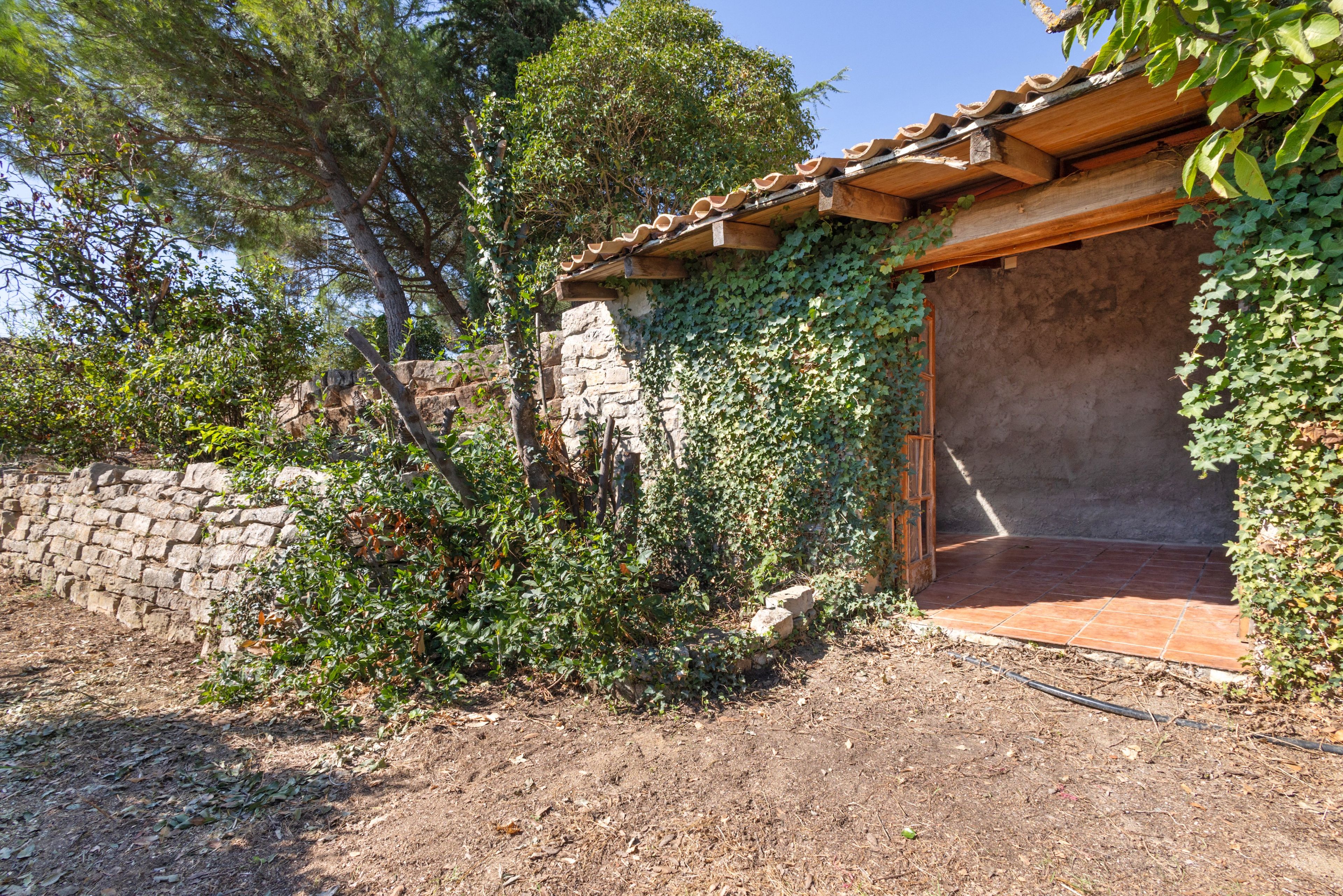 Un bâtiment rustique avec un mur de pierre et des poutres en bois, entouré de verdure et d’arbres.