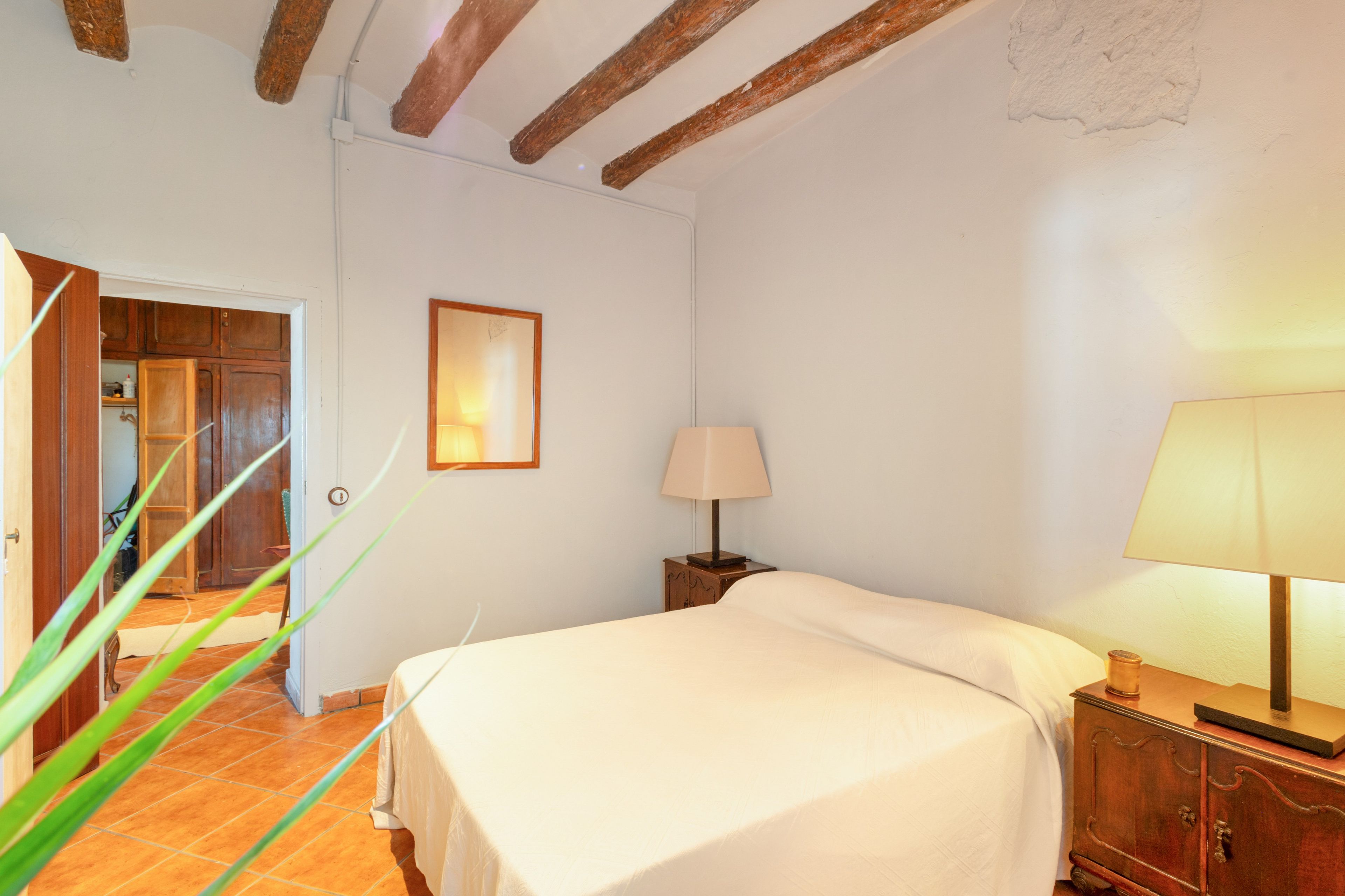 Een slaapkamer met een bed, een nachtkastje, een lamp, een lijst met een afbeelding aan de muur en een houten deur die naar een andere kamer leidt.