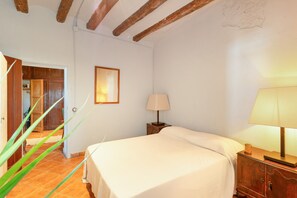 Un dormitorio con cama, mesita de noche, lámpara, un cuadro enmarcado en la pared y una puerta de madera que da a otra habitación.