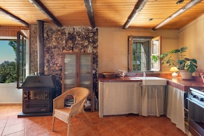 A rustic kitchen with a stone wall, a wooden ceiling, a wicker chair, a stove, and a window with a view.