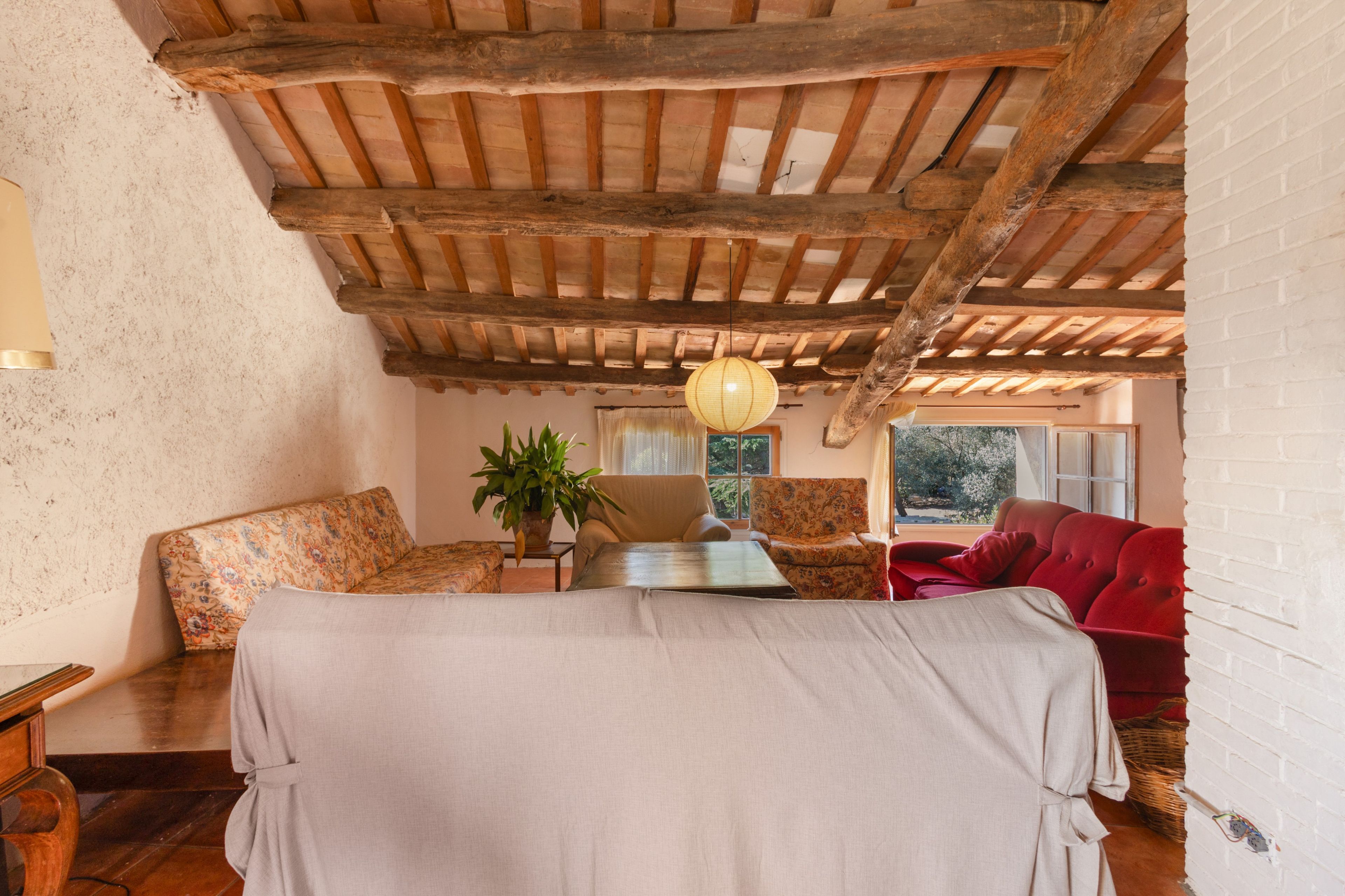 A room with a wooden ceiling, a bed with a white bedspread, a red sofa, and a chair with a floral pattern.