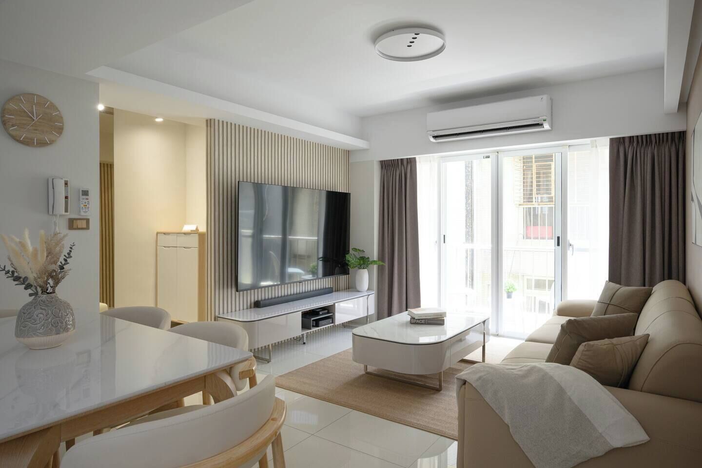 A modern living room with a flat-screen TV, a dining area with a white table and chairs, and a sofa.