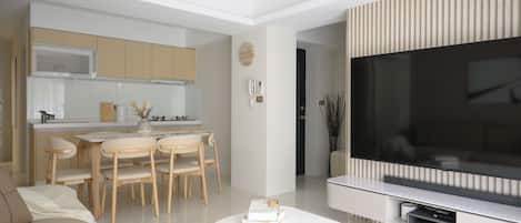 A modern living room with a flat-screen TV, a dining area with wooden chairs, and a kitchen with a microwave.