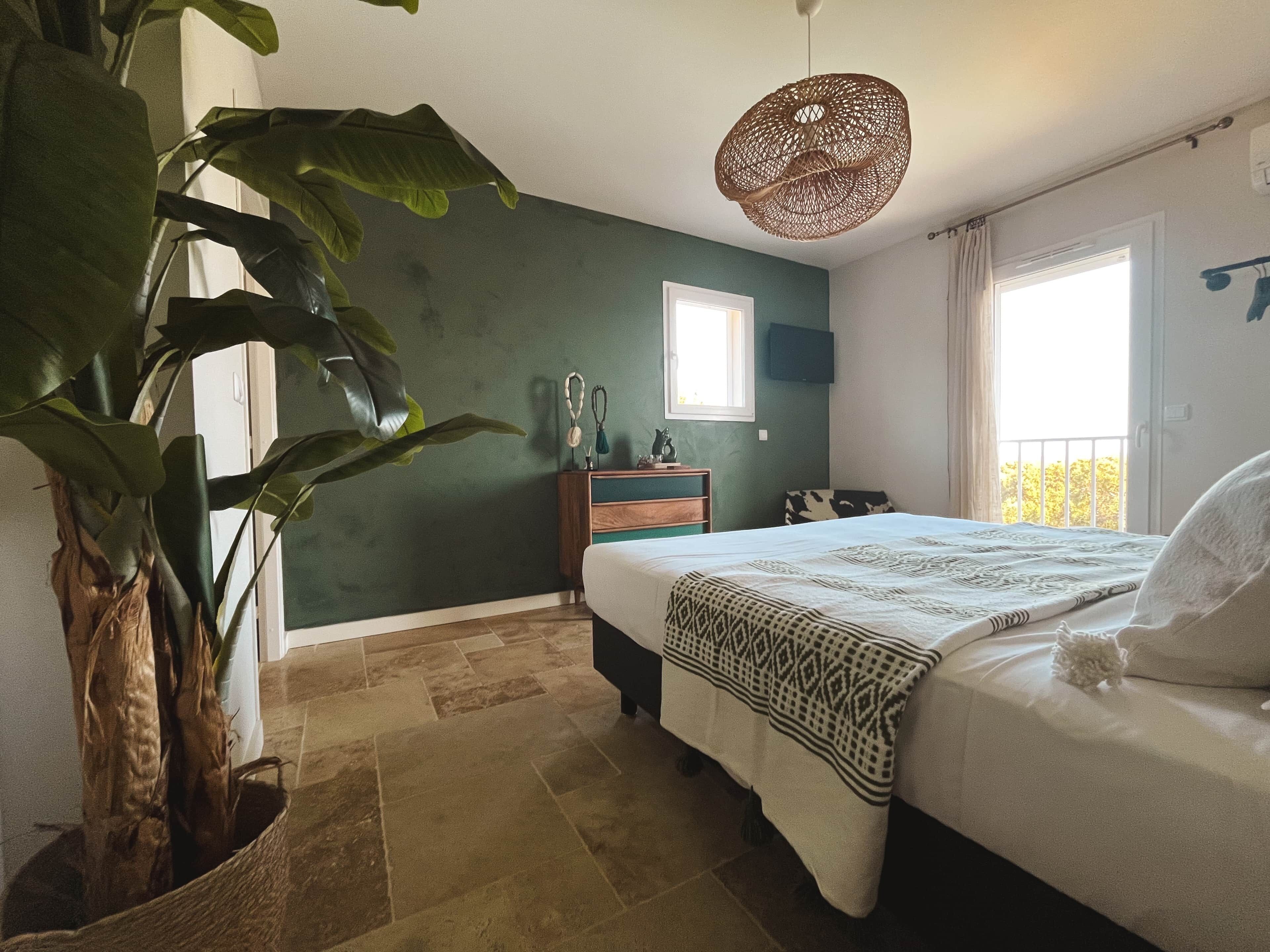A bedroom with a bed, a bedside table, a hanging light fixture, a window, and a plant.