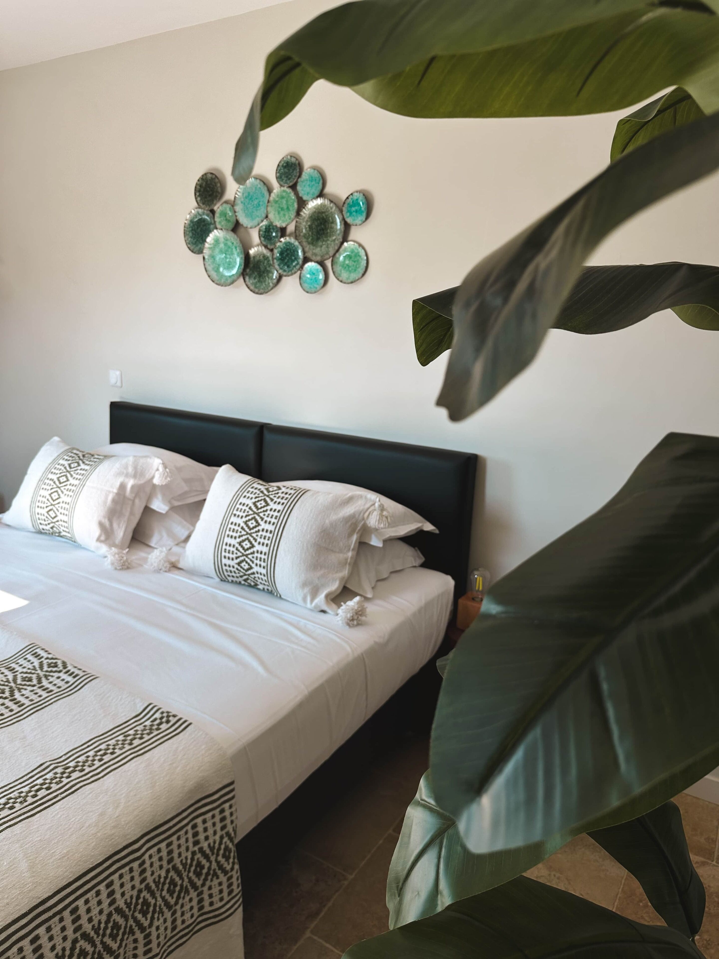 Un lit avec une literie à motifs, un objet décoratif fixé au mur et de grandes feuilles vertes au premier plan.