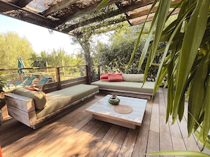Ett trädäck med en soffa och en bänk, ett litet bord med en vas och en pergola med klätterväxter.