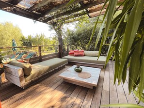 Ett trädäck med en soffa och en bänk, ett litet bord med en vas och en pergola med klätterväxter.