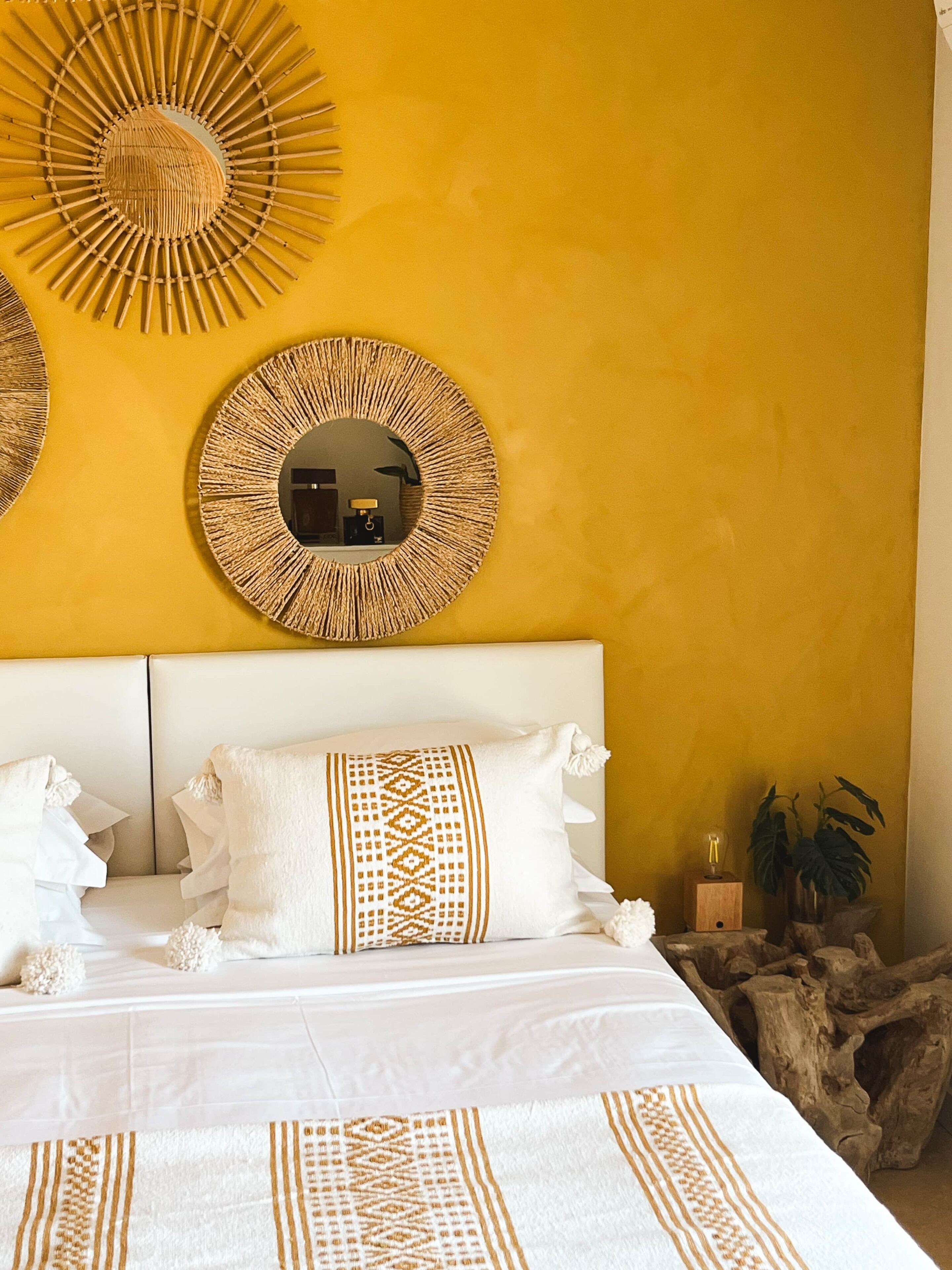 A bed with white bedding and decorative pillows, a round mirror with a woven frame, and a wooden tree stump decoration on the floor.