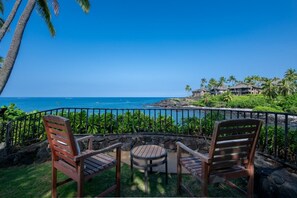 A patio with two wooden chairs and a small table, overlooking a coastal area with a clear view of the ocean and a distant shoreline with buildings.