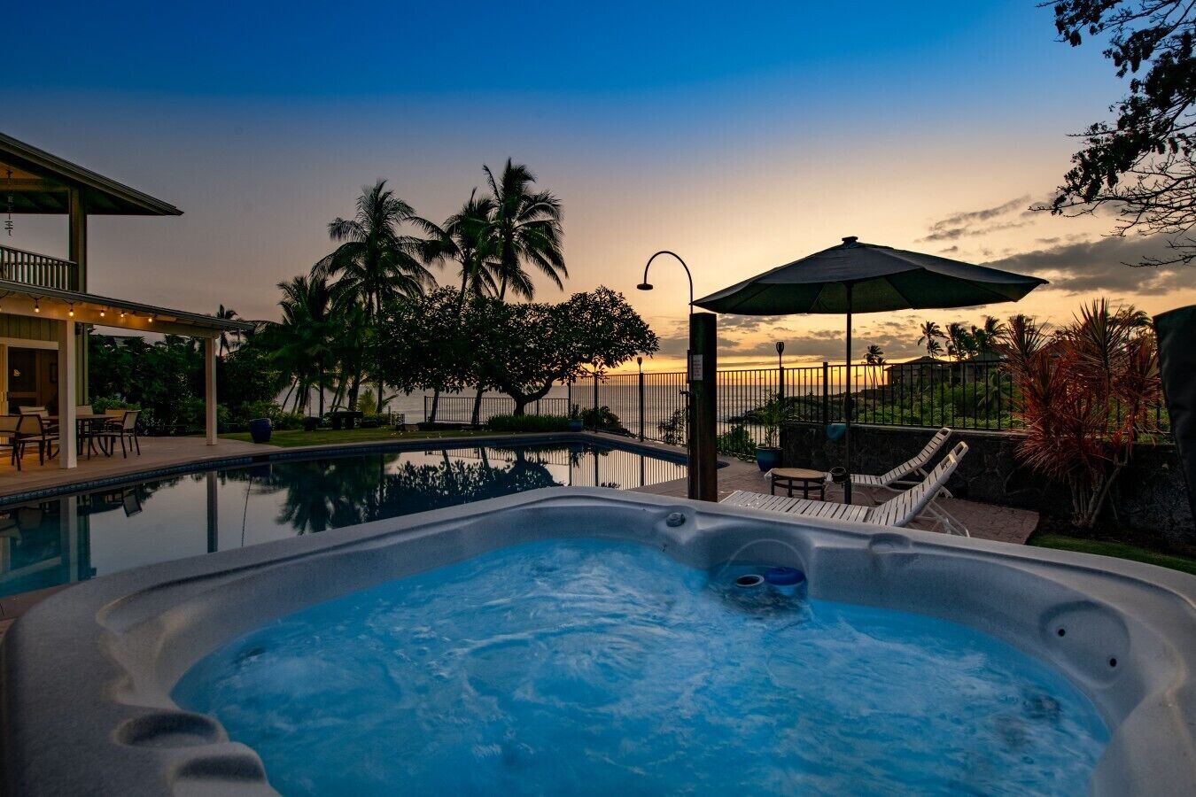 A hot tub with a view of the ocean at sunset, a covered patio area, and a pool.
