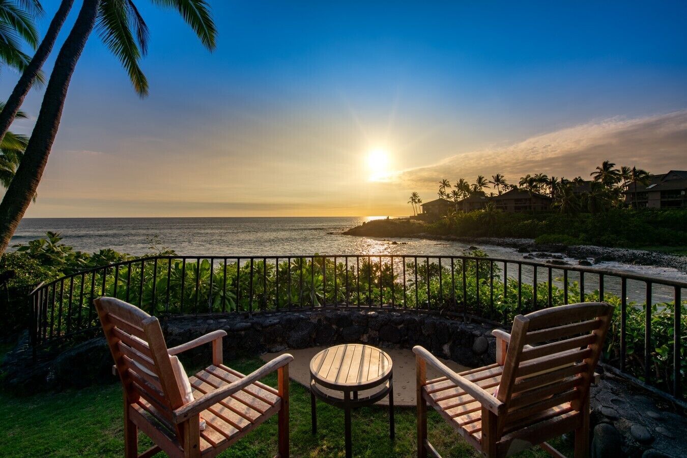 A sunset view from a balcony with two wooden chairs and a small round table, overlooking a beach with palm trees and a rocky shoreline.