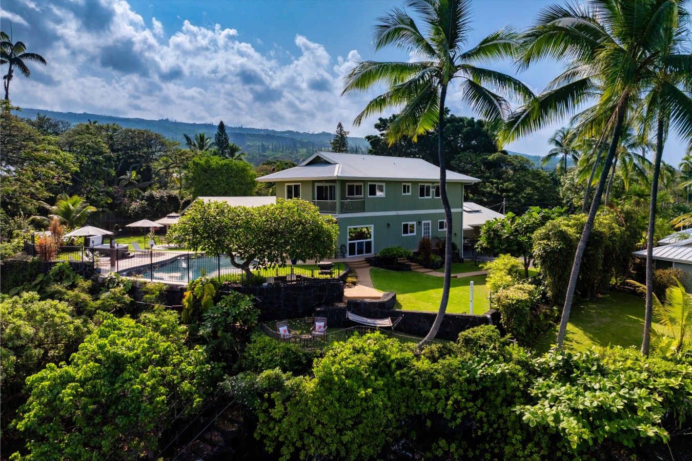 A two-story house with a swimming pool, surrounded by lush greenery and palm trees.