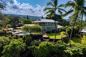 A two-story house with a swimming pool, surrounded by lush greenery and palm trees.