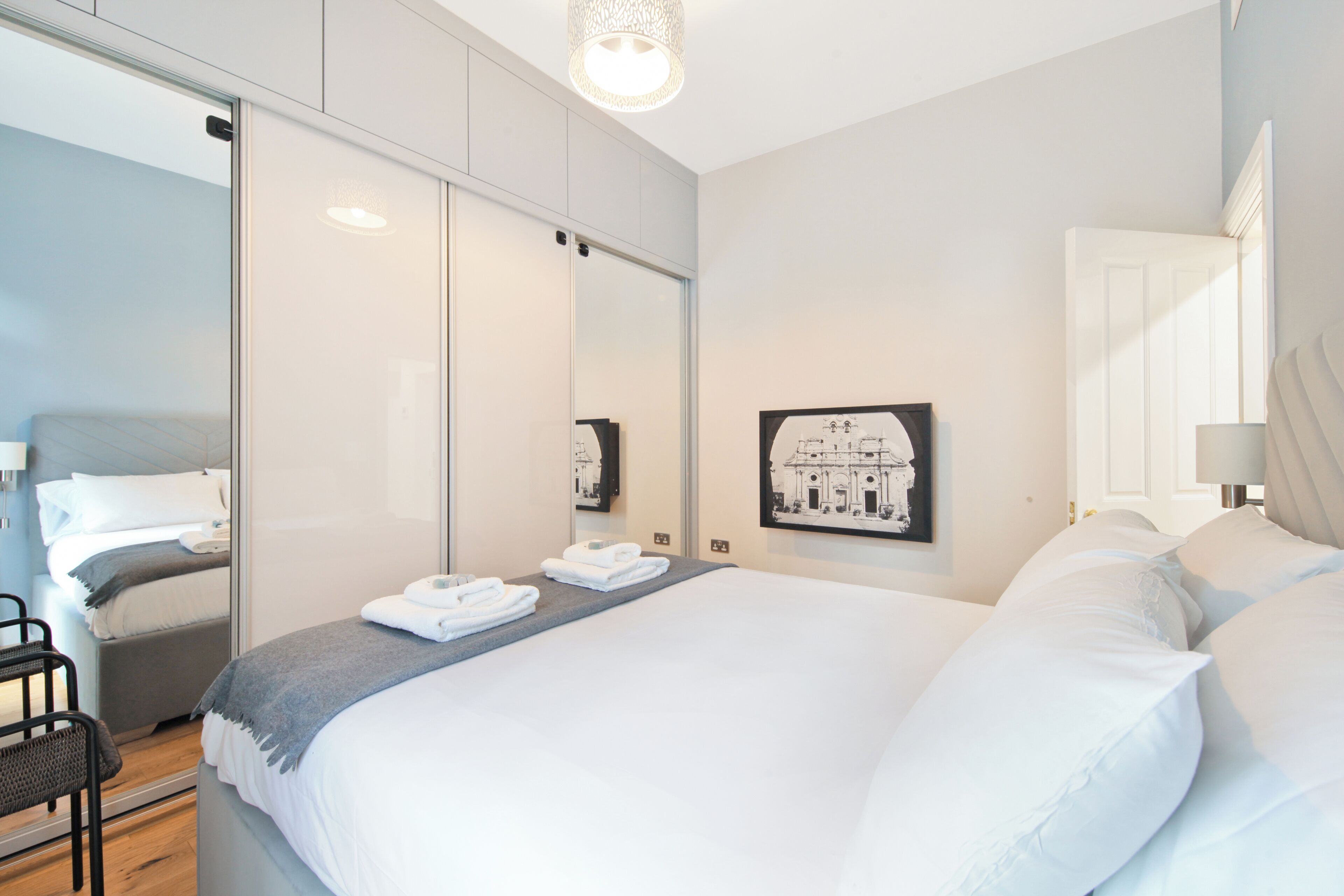 A modern hotel room with a large bed, a bedside table, a chair, and a wall-mounted picture.