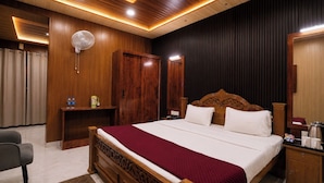 A hotel room with a wooden bed, a bedside table, a fan, and a wardrobe. - The Gautam Resort (Varanasi)