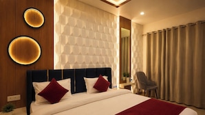 A modern hotel room with a large bed, a headboard with built-in lighting, a wall-mounted mirror, and a seating area with a chair. - The Gautam Resort (Varanasi)