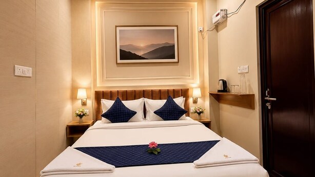 A hotel room with a bed, bedside lamps, a wall-mounted air conditioner, a framed picture, and a door. - Hotel Tapovan Inn (Varanasi)