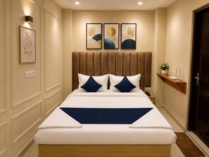 A hotel room with a large bed, a headboard, a nightstand, a wall-mounted shelf with decorative items, and framed artwork on the wall. - Hotel Tapovan Inn (Varanasi)