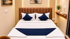 A modern hotel room with a large bed, a headboard with vertical panels, a wall-mounted air conditioner, a bedside table with a plant, and a framed picture on the wall. - Hotel Tapovan Inn (Varanasi)