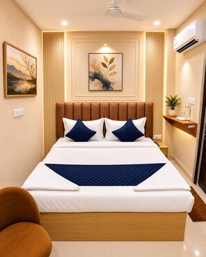 A modern hotel room with a large bed, a headboard with vertical panels, a wall-mounted air conditioner, a bedside table with a plant, and a framed picture on the wall.