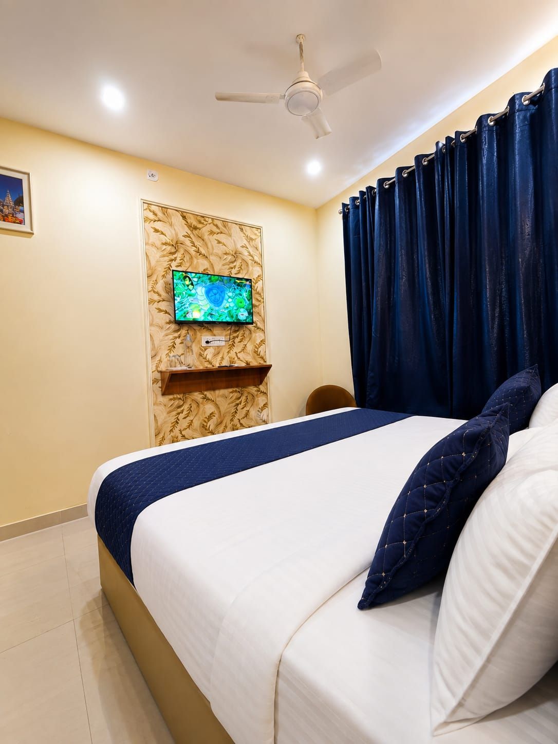 A hotel room with a bed, a ceiling fan, a mounted television, and dark curtains.