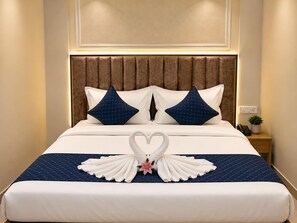 Una habitación de hotel con una cama, dos almohadas, una mesita de noche con un teléfono y una planta, y una pared con un interruptor de luz.