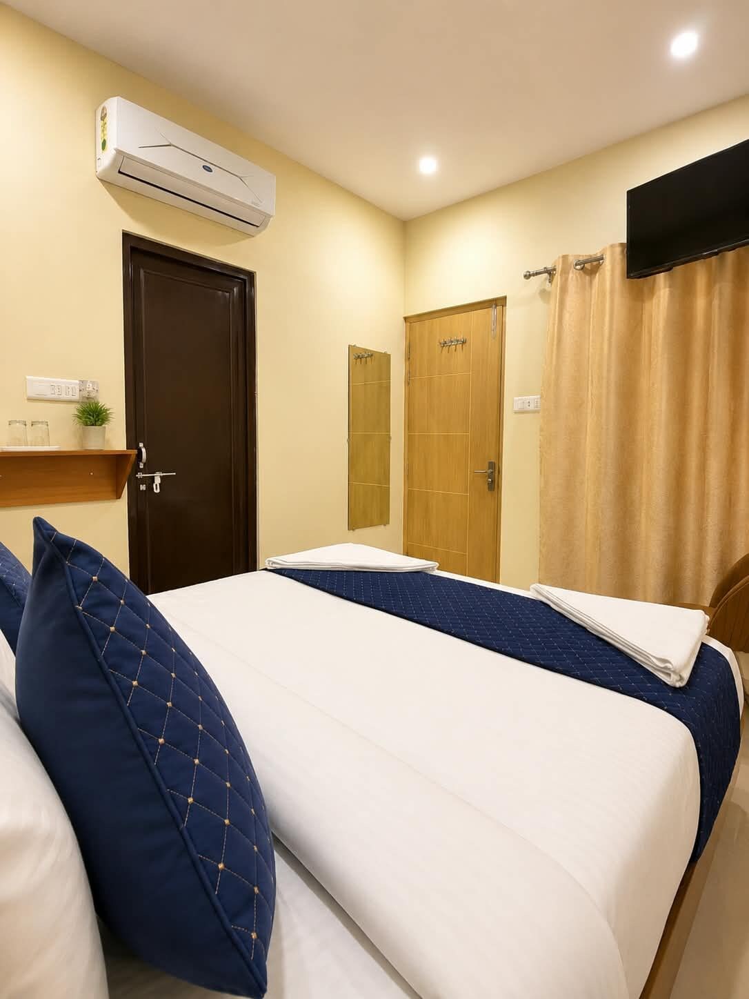 A hotel room with a bed, two doors, an air conditioning unit, a shelf with items, and a curtain.