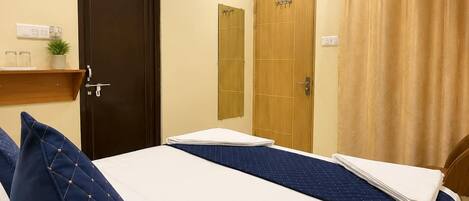 A hotel room with a bed, two doors, an air conditioning unit, a shelf with items, and a curtain.