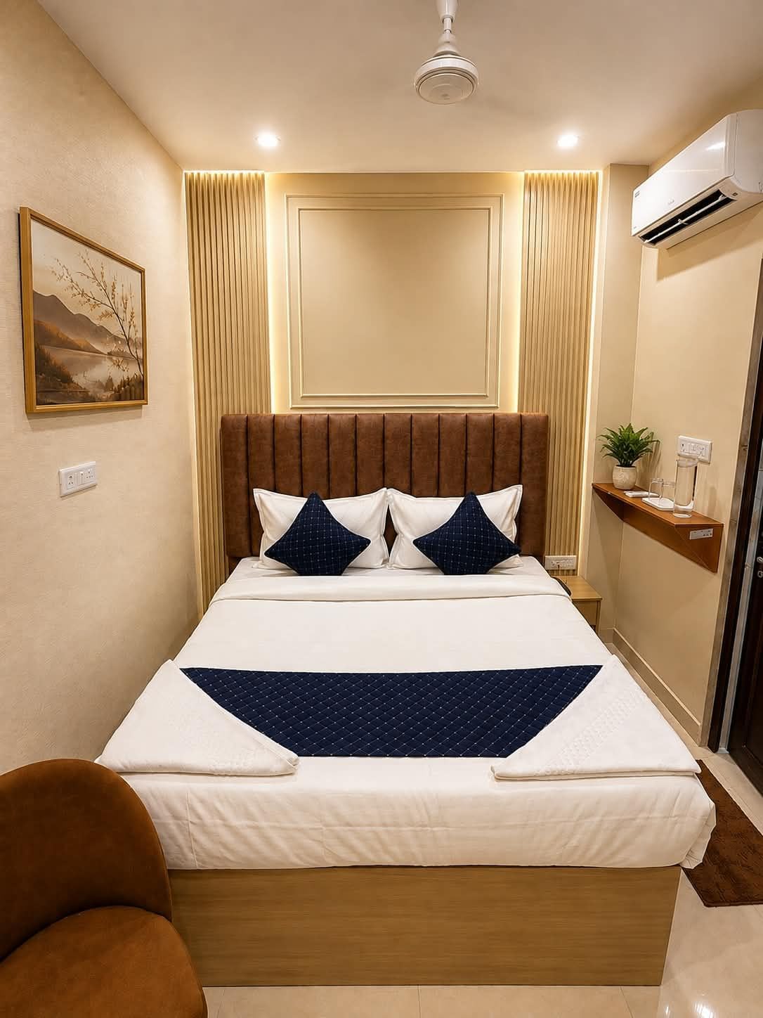 A hotel room with a large bed, a headboard, a wall-mounted air conditioner, a bedside table with a plant, and a painting on the wall.