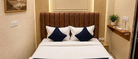 A hotel room with a large bed, a headboard, a wall-mounted air conditioner, a bedside table with a plant, and a painting on the wall.