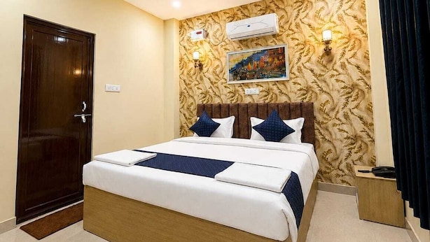 A hotel room with a large bed, a wooden headboard, a wall-mounted air conditioner, and a painting. - Hotel Tapovan Inn (Varanasi)