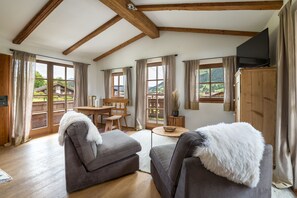 Ein gemütlicher Raum mit Holzbalken, einem Holztisch, zwei grauen Sofas mit weißen Decken und großen Fenstern mit Blick auf ein Haus und Grünflächen.
