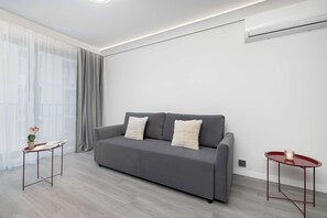 A modern living room with a grey sofa, two white cushions, a small side table, a potted plant, and a red side table. - Comfortable Apartment with Parking Space | 2 Bedrooms | City Centre (Lublin)
