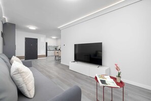 A modern living room with a grey sofa, a flat-screen TV mounted on a white console, and a small table with a magazine and a potted plant. - Comfortable Apartment with Parking Space | 2 Bedrooms | City Centre (Lublin)