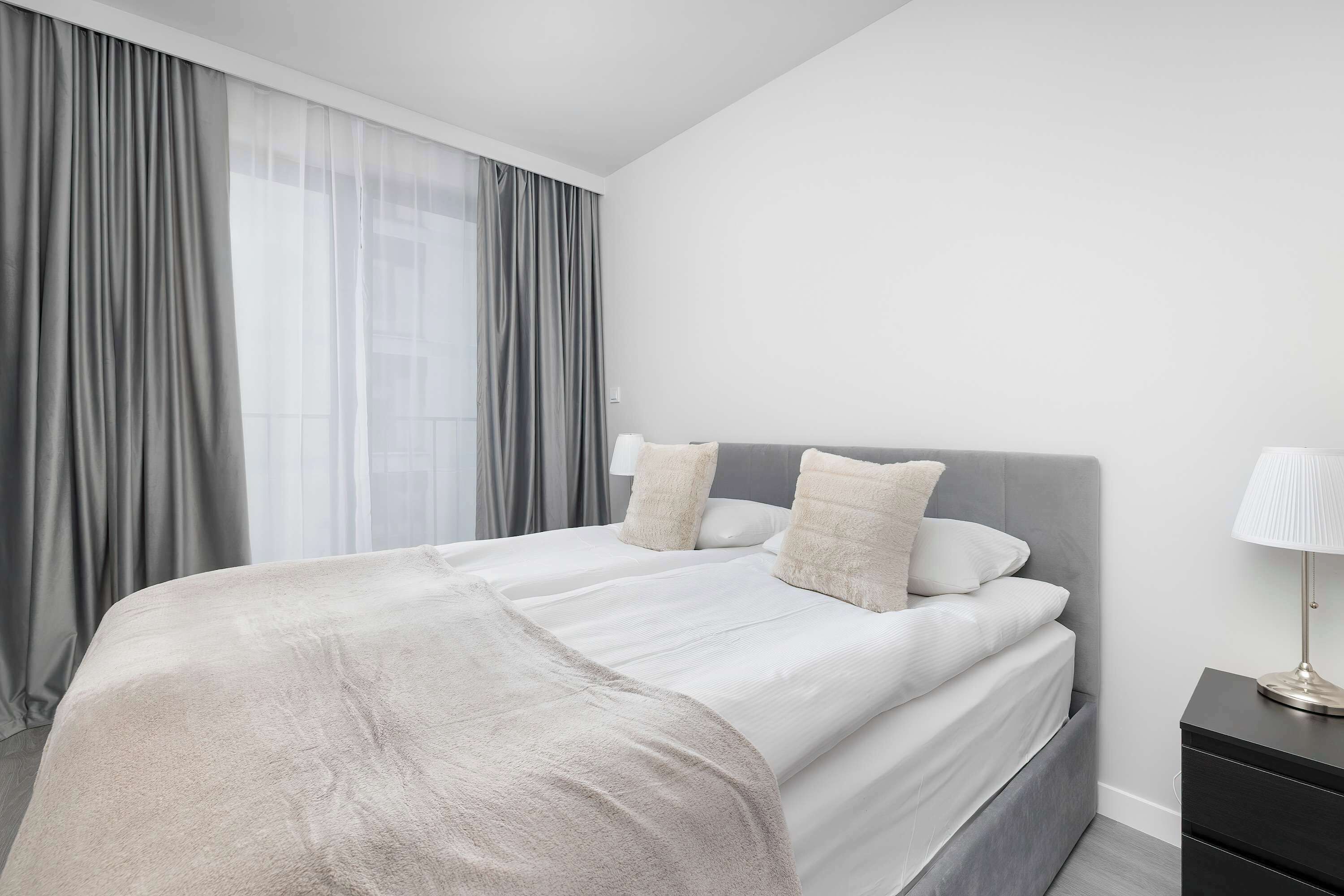 A bright bedroom with a large double bed, white bedding, and soft grey curtains framing the window. A small bedside table with a lamp completes the simple, elegant arrangement.