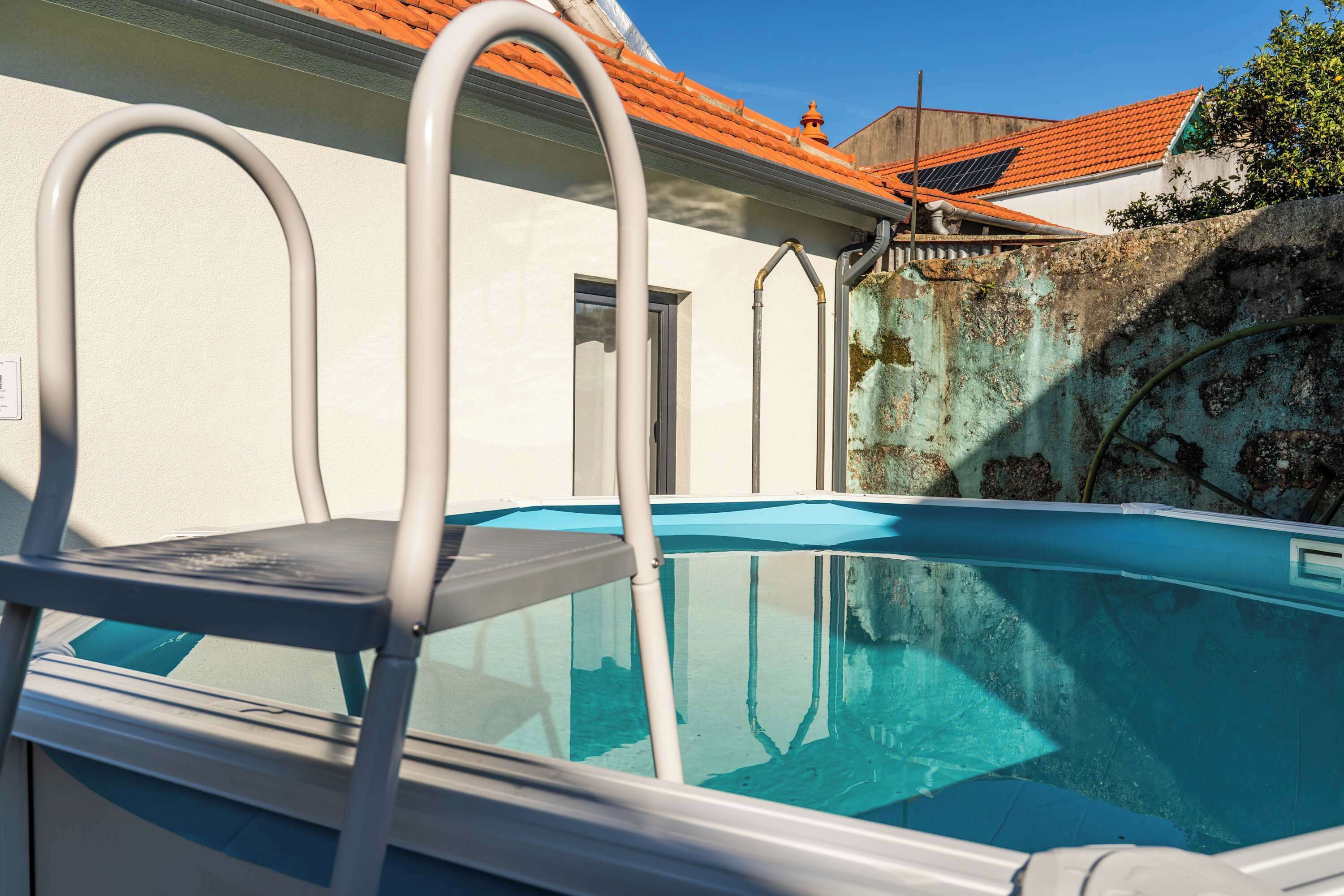 A swimming pool with a metal ladder, a white wall, and a red-tiled roof.