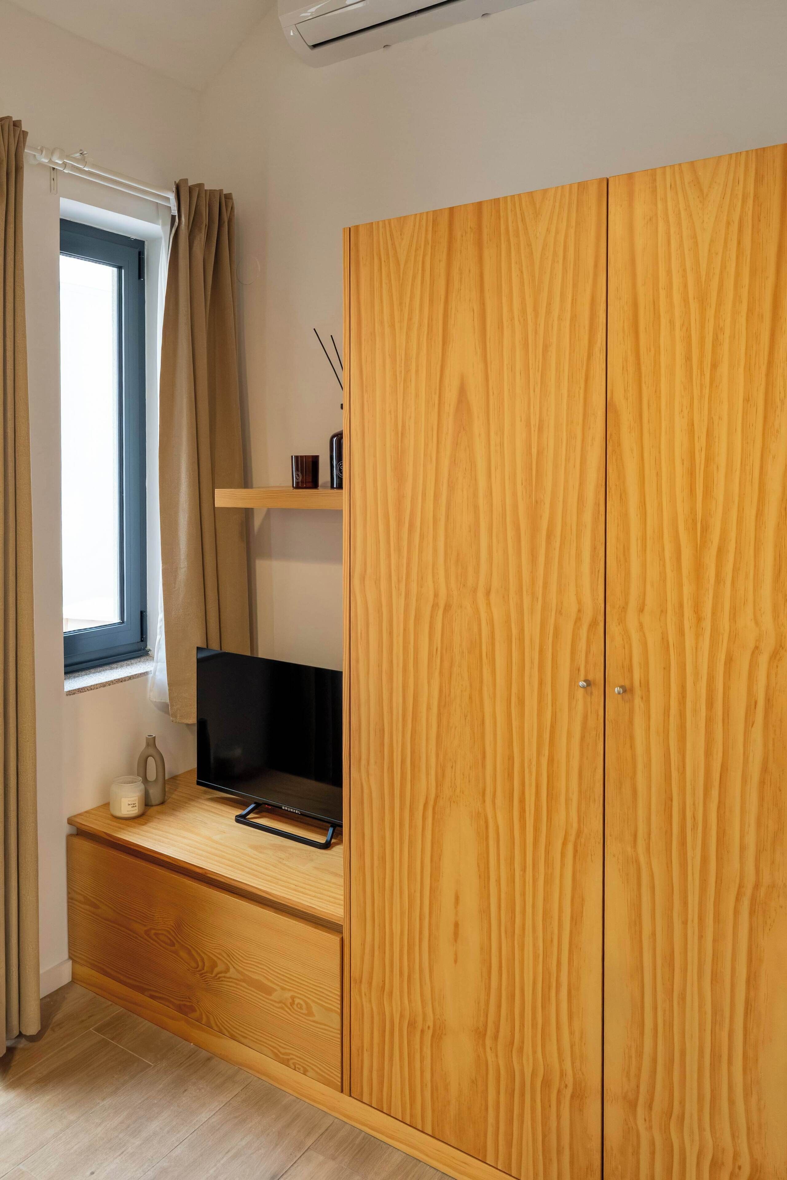 A room with a wooden wardrobe, a small desk with a laptop, a window with curtains, and a shelf with decorative items.