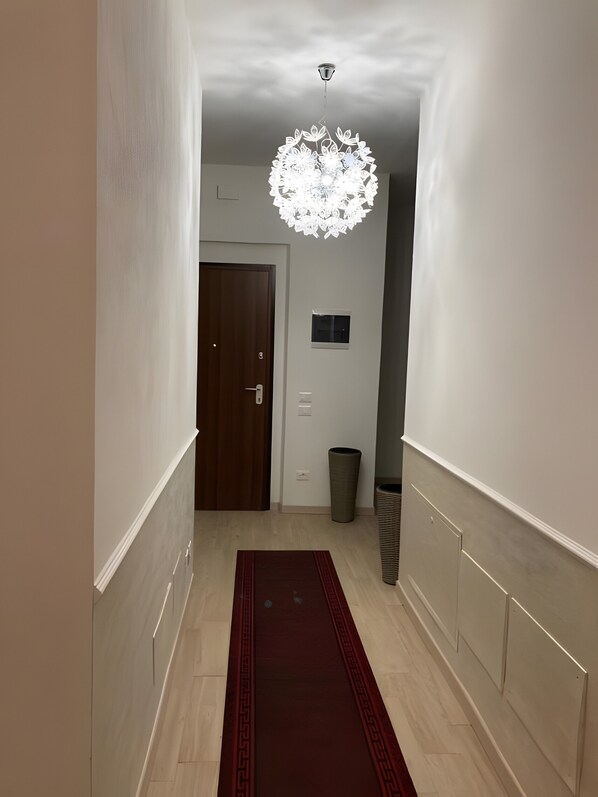 A hallway with a wooden door, a trash bin, a wall-mounted light fixture, and a red rug.