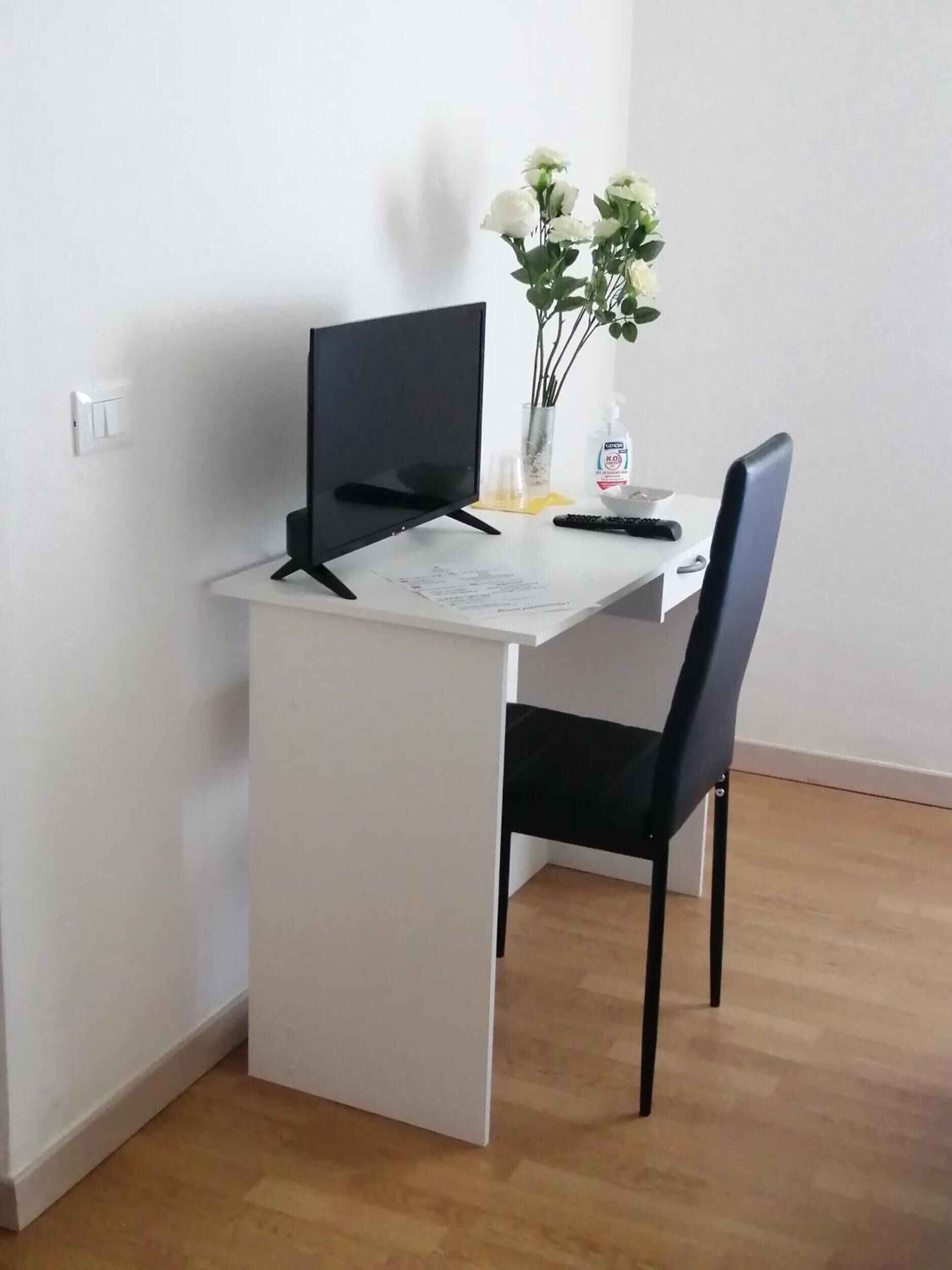 Een klein wit bureau met een zwarte stoel, een afstandsbediening, een vaas met witte bloemen en een fles handdesinfectiemiddel.