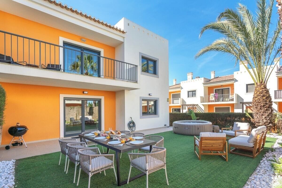 A two-story residential building with a balcony, a dining area with a table and chairs, and a hot tub.