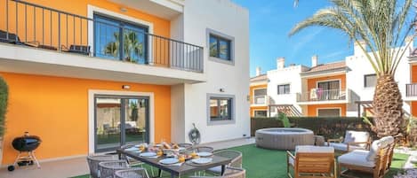 A two-story residential building with a balcony, a dining area with a table and chairs, and a hot tub.