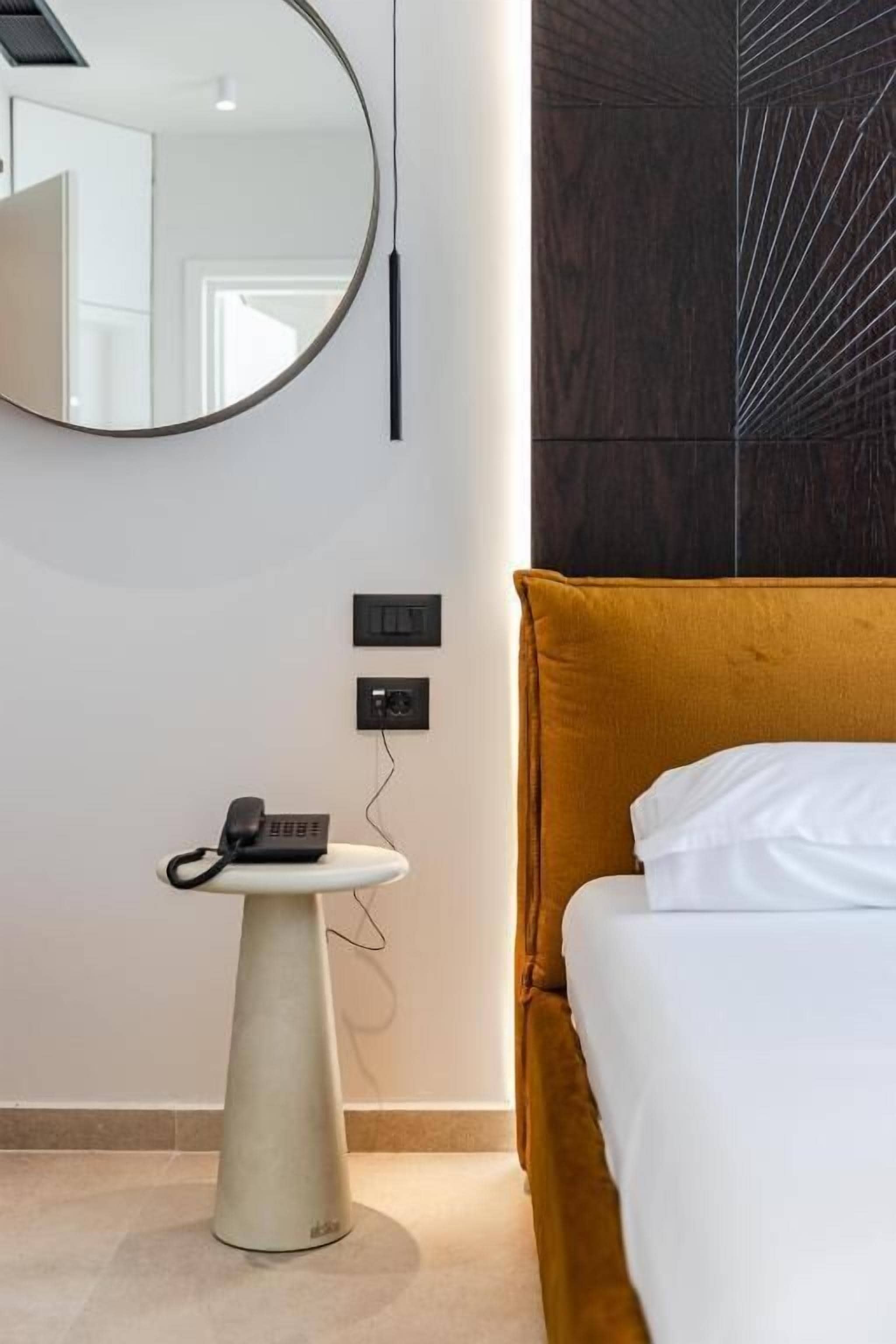 Ein Hotelzimmer mit einem Bett, einem runden Spiegel, einem kleinen Tisch mit Telefon und einer Wandleuchte.