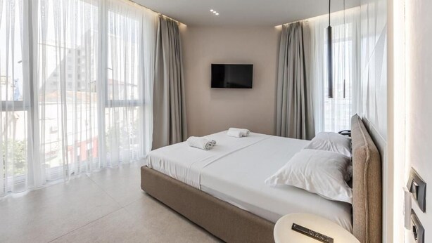 Una habitación de hotel moderna con una cama grande, un televisor de pantalla plana y vistas de la ciudad a través de cortinas translúcidas.
