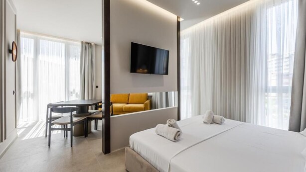 Una habitación de hotel con una cama, un escritorio, una silla, un televisor montado en la pared y un amplio ventanal con cortinas.