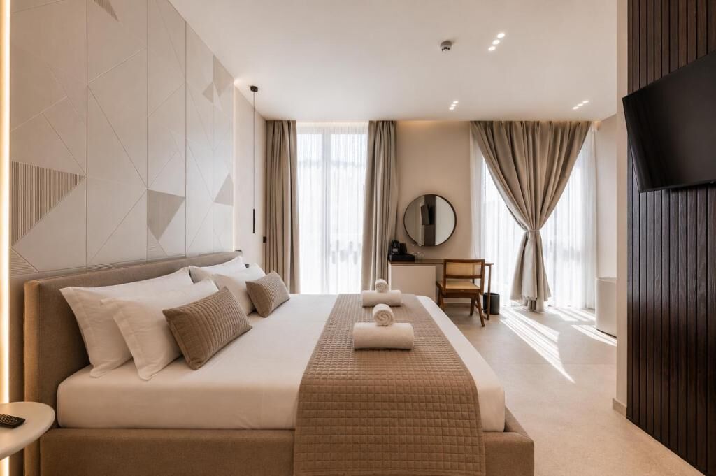 Habitación de hotel moderna con una cama grande, un escritorio, un televisor de pantalla plana y un amplio ventanal con cortinas.